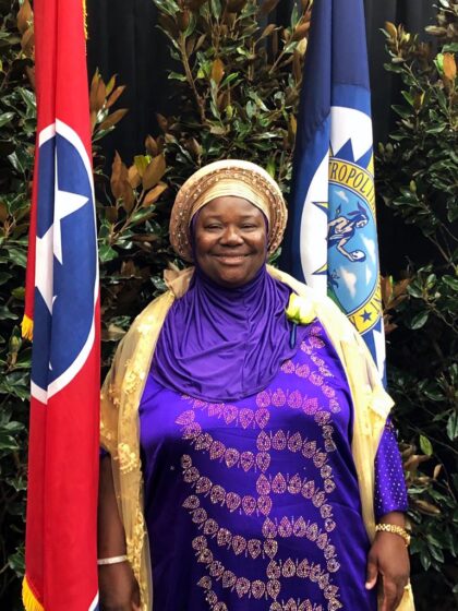 A Black woman with a purple dress and a headscarf stands beside the TN flag